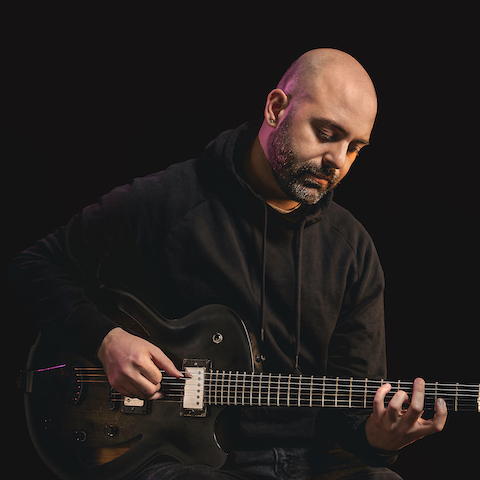 Caio Afiune playing guitar in a black hoodie against a black backdrop