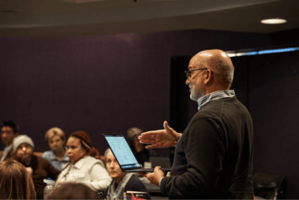 A picture of someone with a laptop speaking to a crowd. 