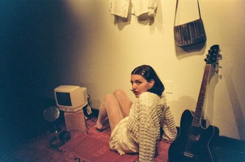 Analise sits in a dimly lit room. She wears all white and looks back at the camera.  