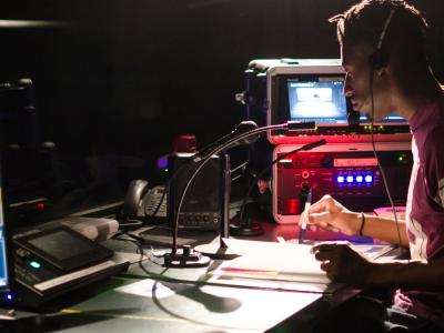 Man with headset seated at a work station surrounded by recording devices and other audio equipment