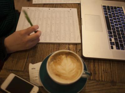 Person writing on blank sheet music at a desk with coffee and a laptop