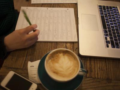 Person writing on blank sheet music at a desk with coffee and a laptop