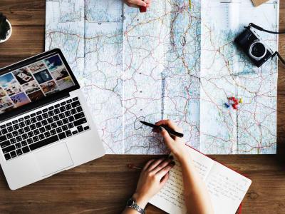 Person marking up a large map on a table with a laptop, camera and notebook