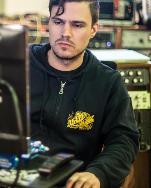 Benny Grotto sitting and looking at a computer in his recording studio, surrounded by gear