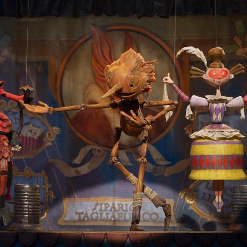 A film still of a wooden Pinocchio on stage in between a devil puppet to the left and a woman puppet to the right
