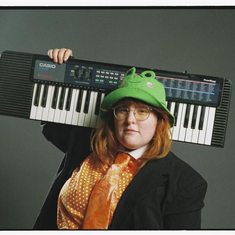 corook wearing a frog hat, holding a keyboard on their shoulder