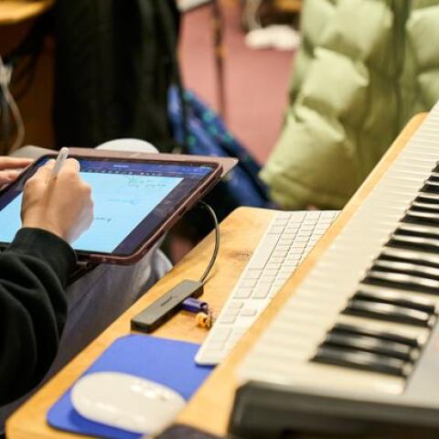 A person's hands writing on a digital tablet next to a MIDI keyboard workstation