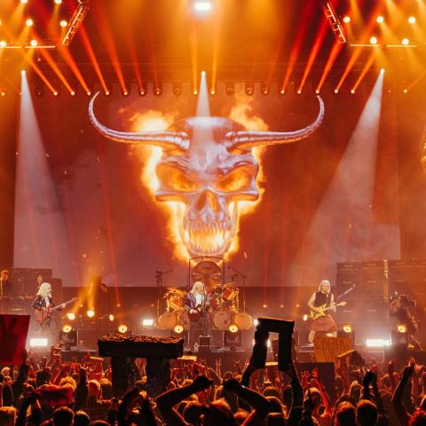 Spinal Tap on stage from a crowd perspective, with fans holding up miniature stonehenges and a giant flaming skull behind the band