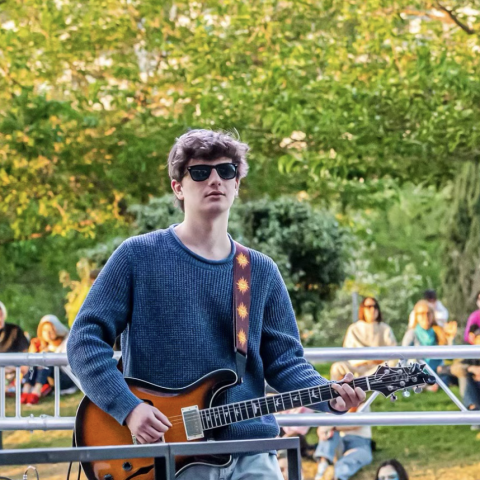 Jesse stands on an outdoor stage with a guitar. He's wearing a blue sweater, light wash jeans, and glasses. 