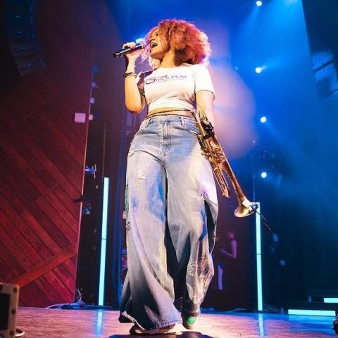 Lady Satch stands on stage in blue jeans and a white tank top, holding her trumpet and a microphone 