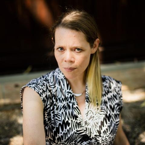 Caroline Davis looks directly at the camera. They have straight blond hair pulled in a side ponytail and a black and white patterned shirt