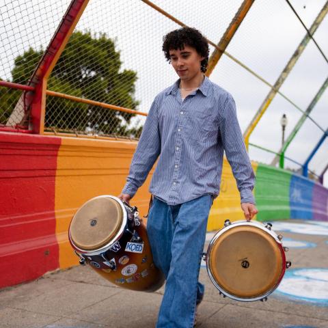 Ceferino Vergara alking across a colorful bridge while carrying two hand drums, with a rainbow-painted wall and chain-link fence in the background.
