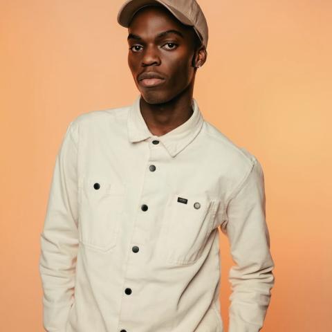 Mitchel Kedi of wearing a light button-up shirt and a baseball cap, standing against a warm orange background with a neutral expression.