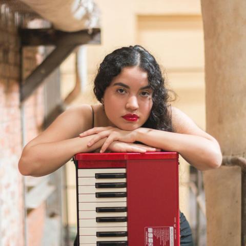 Magalí Benítez with curly hair resting her arms on a red keyboard, looking directly at the camera in an indoor setting with brick and concrete walls.