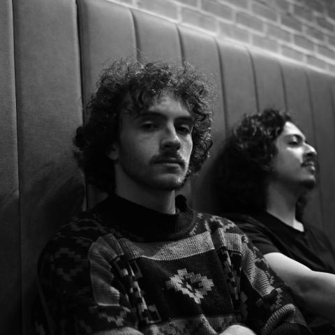 Black-and-white photo of Liam Backhoff sitting against a padded wall, one facing the camera with a calm expression while the other leans back with eyes closed.