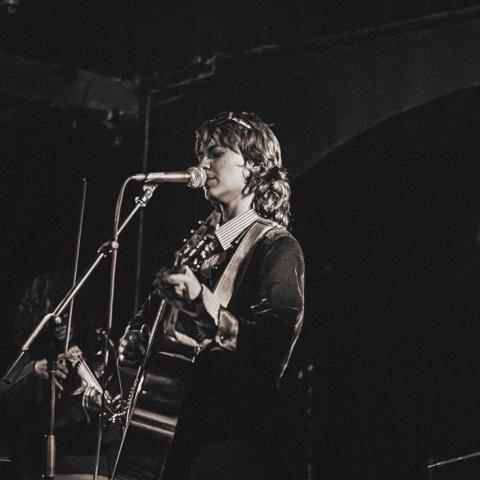     Black-and-white photo of Marianthi Olympia singing into a microphone while playing guitar on stage, with another musician partially visible in the background.