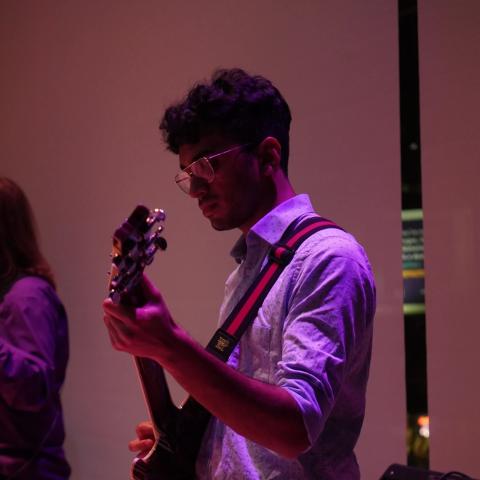 Devesh Iyer Trio wearing glasses playing an electric guitar under purple lighting, focused on the instrument during a live performance.