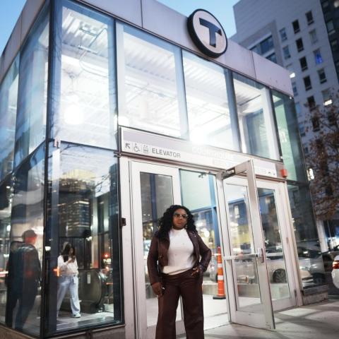 Vanessa Gracious Gift standing confidently outside a glass transit station entrance at dusk, with city buildings and street activity in the background.