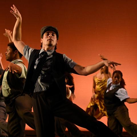 Dancers on stage in front of a n ornage background. Dancer in foreground has left hand lifted towards the ceiling with the right hands horizontal towards the right side.