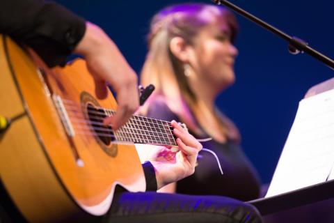 Closeup of a hand playing a nylon stringed guitar while a vocalist is in the background