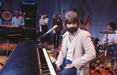 A young Michael McDonald sitting on stage at a piano with his band in the background