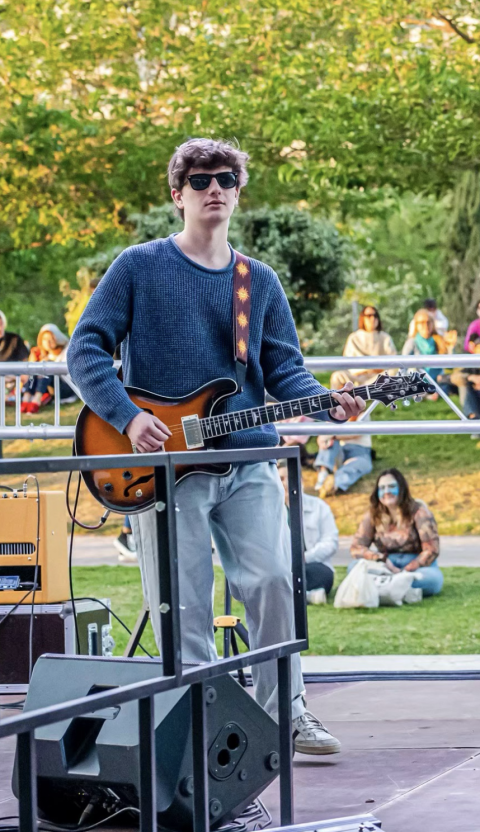 Jesse stands on an outdoor stage with a guitar. He's wearing a blue sweater, light wash jeans, and glasses. 