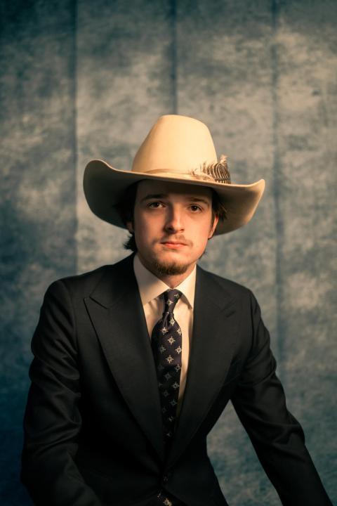 Nico Welsh wearing a suit and stetson hat posing against a gray backdrop