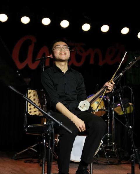 Yu sitting on stage holding a traditional string instrument, smiling under bright stage lights with drums in the background.