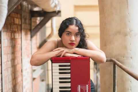 Magalí Benítez with curly hair resting her arms on a red keyboard, looking directly at the camera in an indoor setting with brick and concrete walls.