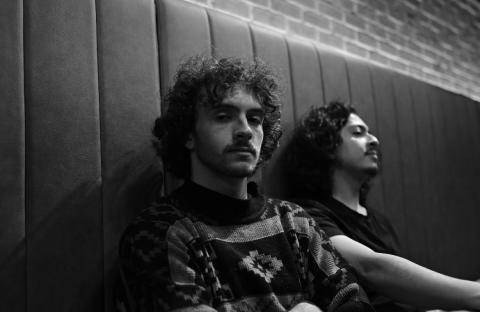 Black-and-white photo of Liam Backhoff sitting against a padded wall, one facing the camera with a calm expression while the other leans back with eyes closed.