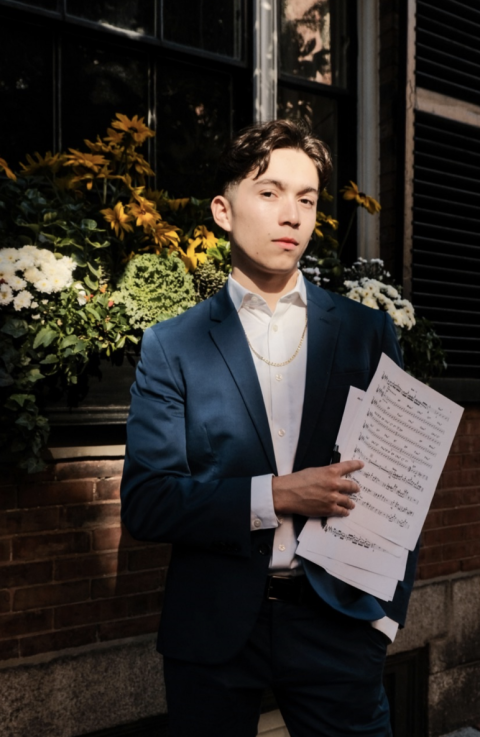 Montaño Quartet in a suit holding sheet music, standing outdoors in front of flowers and a window, looking directly at the camera.