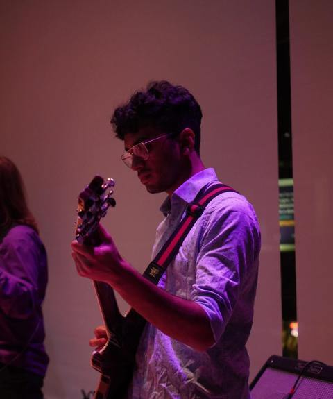 Devesh Iyer Trio wearing glasses playing an electric guitar under purple lighting, focused on the instrument during a live performance.