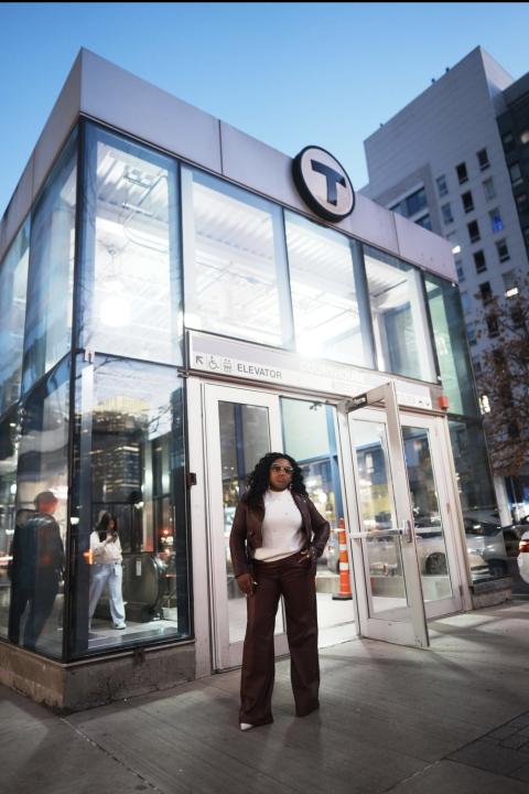 Vanessa Gracious Gift standing confidently outside a glass transit station entrance at dusk, with city buildings and street activity in the background.
