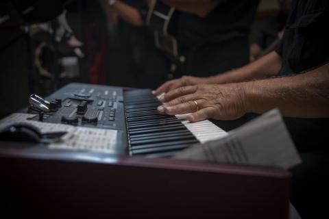 A close-up shot shows a person's hands playing an electronic keyboard during a live performance. In the dimly lit background, other musicians can be softly seen.