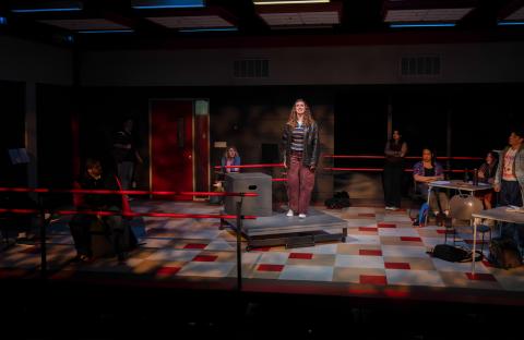 person singing on stage with a spotlight and small podium beneath their feet. The floor is checkered.