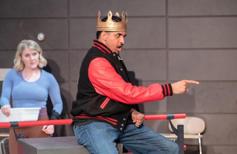 person in a varisty jacket (black adn red) wearing a crown singing and pointing