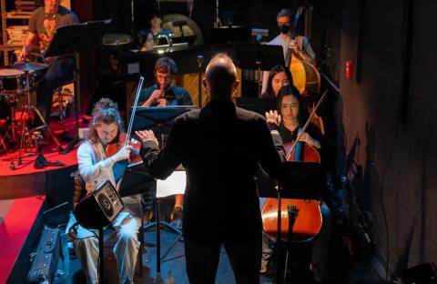 Pit orchestra of 'Time to Act' playing: back of conductor and string section are visible.