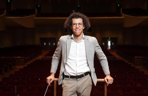Jonathon Heyward smiling in a gray suit on a conducting podium in a concert hall.