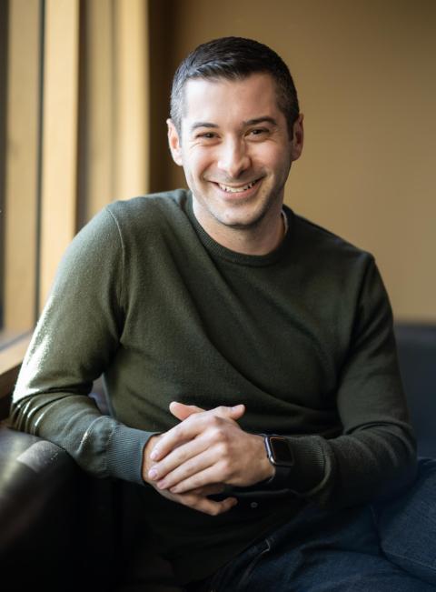 Head-and-shoulders portrait of a smiling person wearing a dark green sweater, seated indoors and facing the camera.